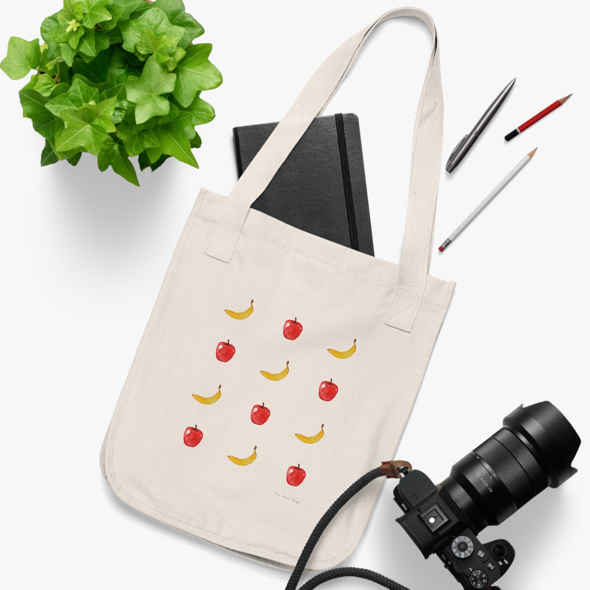 Beige tote bag with fruit pattern on a white surface with a plant, notebook, and camera.
